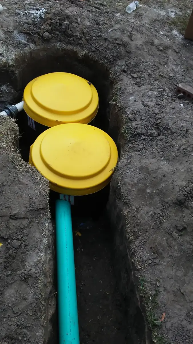 Two yellow-lidded drain basins set in an open trench with green PVC drain pipe leading away