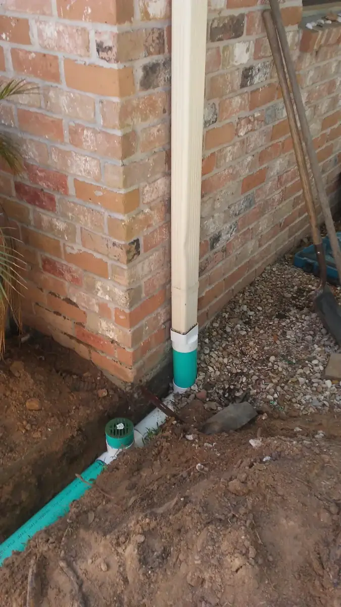 Downspout connected to green underground drain pipe at a brick wall, trench open