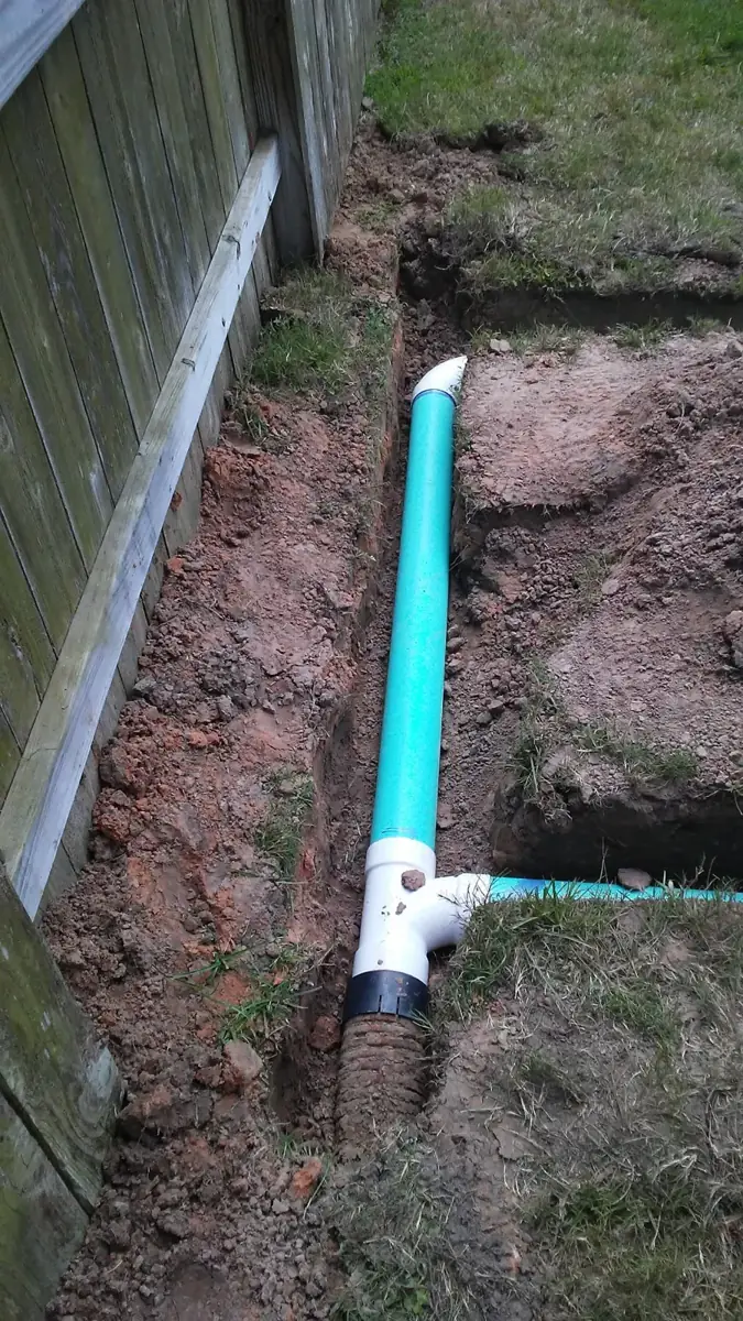 Green drain pipe with a white tee fitting in a clay trench along a wooden fence