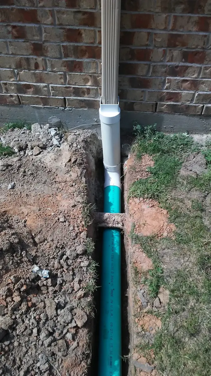 Downspout feeding straight into a green underground pipe in an open trench next to a brick wall